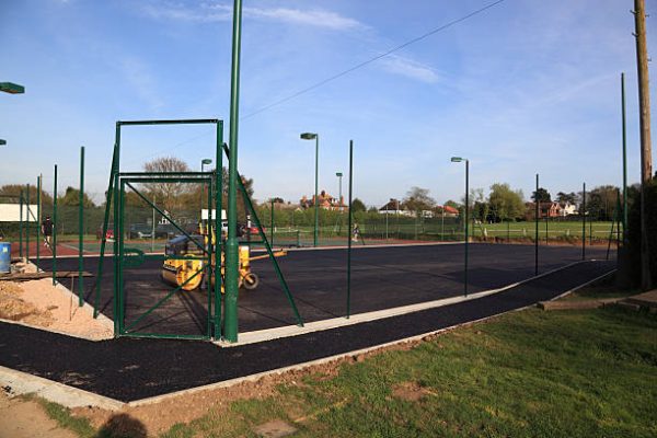 Epping, United Kingdom - April 7, 2011: a new tennis court is under construction in Epping, England. A layer of  asphalt has been laid and compacted by mechanical rollers, which can be seen. A path has also been laid around teh court and the stantions and gate are in place. two tennis players can be seen ni the background on the existing court.